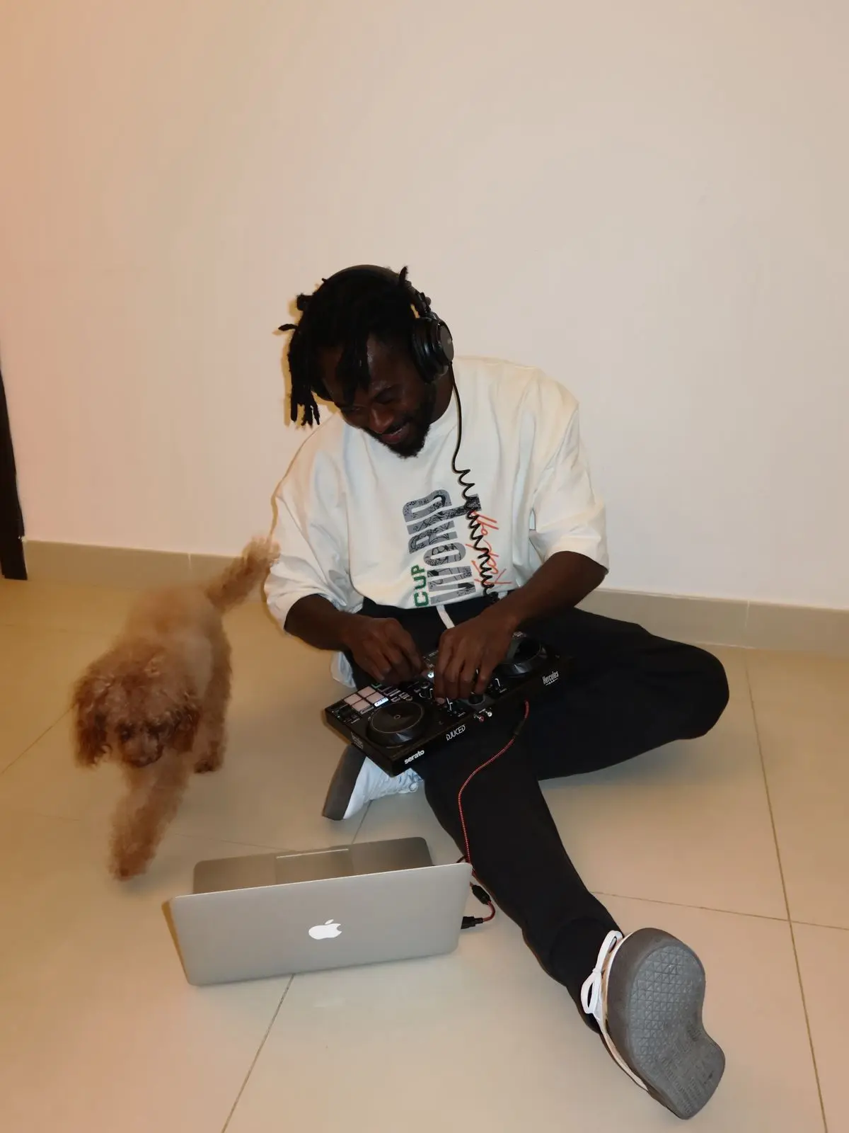 DJ Christerna smiling and mixing music on a portable DJ controller while sitting on the floor, with a small brown poodle walking next to him.